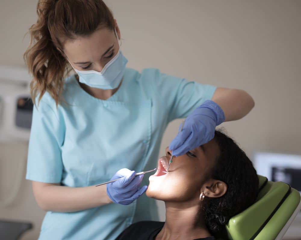 Dental hygienist in blue gloves and mask examining patient's teeth under bright light in clinical setting.